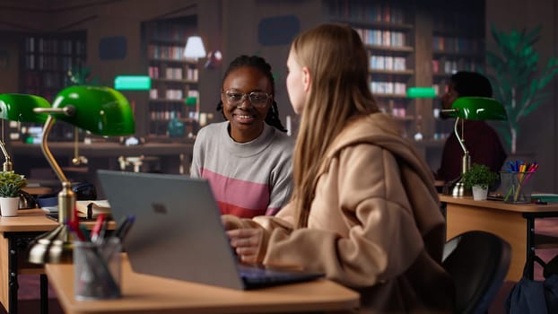 Female student reading and writing detailed notes for an exam, studying political science for upcoming lecture in a library setting at the university. Young adult consults a colleague. Camera A.