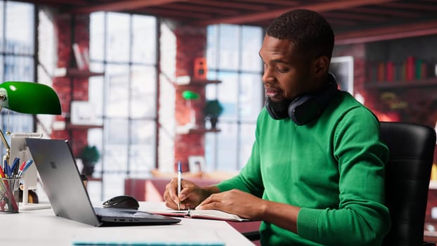 African american male teleworker writing personal notes at his desk in modern loft, remote job. Productive guy sitting at home office, using pen to take notes in his textbook. Camera A.