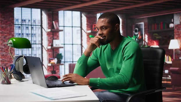 Black man doing remote work feeling uneasy and anxious at desk, shaking his legs and being jittery due to task completion and deadline. Freelancer working under pressure, anxiety. Camera B.