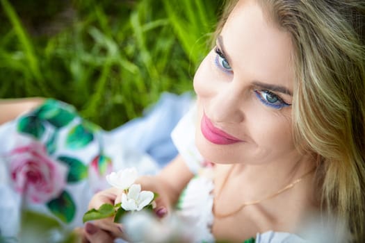 Blonde adult woman smiles warmly while lying in a green field, holding a delicate flower, enjoying a moment of nature and serenity.