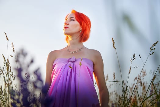 A woman with striking red hair is poised gracefully in a flowing pink dress. She stands amidst tall grass under a colorful sunset sky, creating a serene atmosphere.