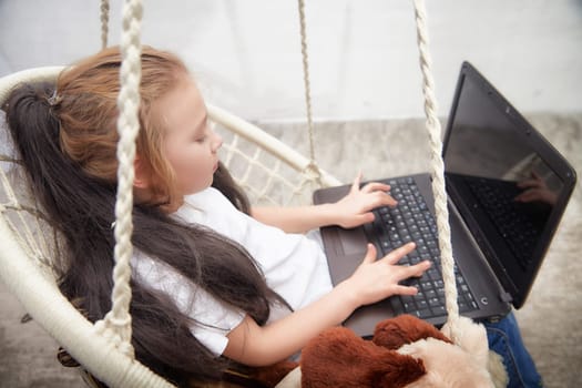 Young child happily types on a laptop while comfortably seated on a swing, engaged in a creative activity during afternoon hours.