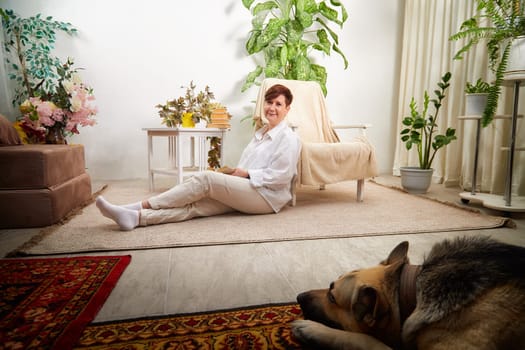 In a cozy room, a middle-aged woman sits relaxed with her German Shepherd dog nearby, surrounded by greenery.