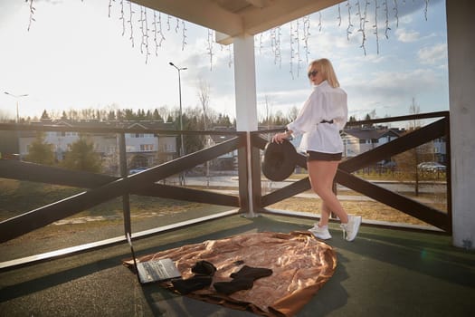 A young woman stands on a balcony, wearing casual clothes and a hat, with a laptop nearby as the sun sets in the background.