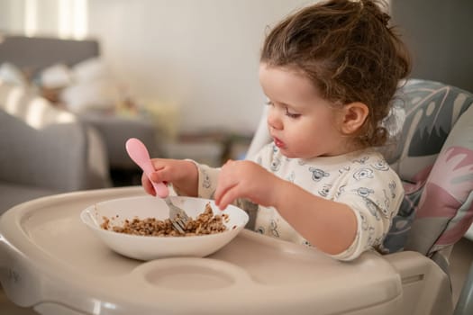 Happy toddler with curls having breakfast in a high chair. Kitchen background with bowl, spoon, and loving family environment.