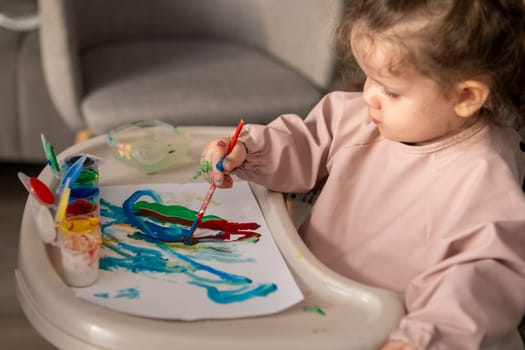 Little girl painting with gouache at a small childrens table. Bright room, toddler engaged in creative activity with colorful paints.