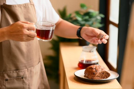 Close up of man in an apron holding a glass cup of black coffee and a piece of bread.