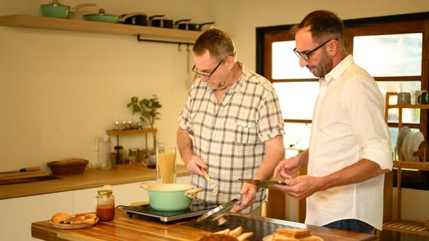 Smiling father and adult son preparing a pasta meal together in a cozy home kitchen. Father day concept.