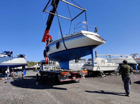 A sailboat lifted by a crane in Suomenoja harbor in Espoo, Finland. A man is holding a rope, guiding the boat as it is suspended in the air by lifting straps