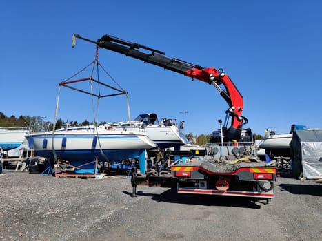 A sailboat lifted by a crane in Suomenoja harbor in Espoo, Finland. A man is holding a rope, guiding the boat as it is suspended in the air by lifting straps