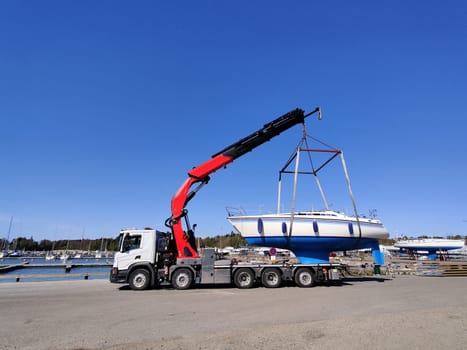 A sailboat lifted by a crane in Suomenoja harbor in Espoo, Finland. A man is holding a rope, guiding the boat as it is suspended in the air by lifting straps