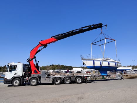 A sailboat lifted by a crane in Suomenoja harbor in Espoo, Finland. A man is holding a rope, guiding the boat as it is suspended in the air by lifting straps