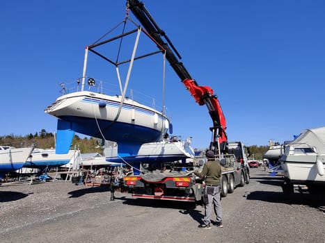 A sailboat lifted by a crane in Suomenoja harbor in Espoo, Finland. A man is holding a rope, guiding the boat as it is suspended in the air by lifting straps