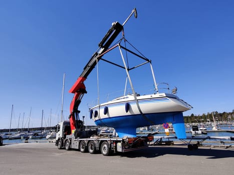 A sailboat is being lifted by a crane in a harbor in Suomenoja, Espoo, Finland. The vessel suspended in the air by heavy lifting straps as it is moved for maintenance or seasonal storage