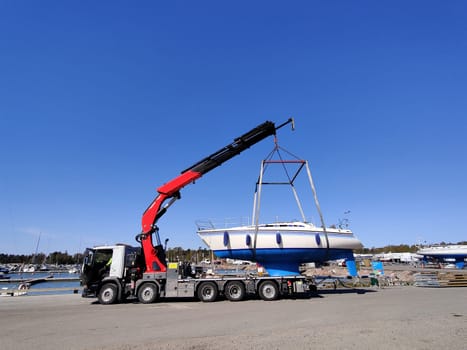 A sailboat is being lifted by a crane in a harbor in Suomenoja, Espoo, Finland. The vessel suspended in the air by heavy lifting straps as it is moved for maintenance or seasonal storage