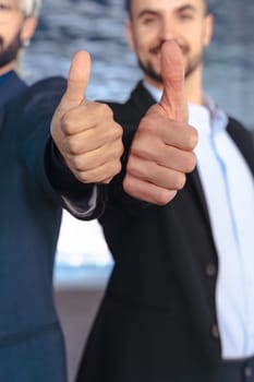 Unrecognizable Two young businessmen in formal suits smiling confidently at the camera, giving thumbs up gesture inside a modern glass office with reflections and stylish interior design elements.
