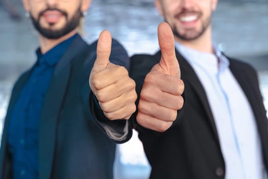 Unrecognizable Two young businessmen in formal suits smiling confidently at the camera, giving thumbs up gesture inside a modern glass office with reflections and stylish interior design elements.