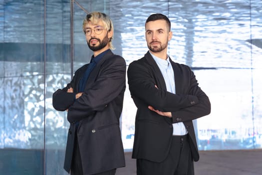 Two confident male professionals in suits standing with crossed arms, serious at the camera inside a modern office. Corporate success, teamwork, and leadership in business environment.