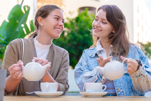 Two Caucasian women sitting at an outdoor cafe, smiling and pouring tea for each other while enjoying a relaxed and cheerful moment of friendship, conversation, and leisure on a sunny day.