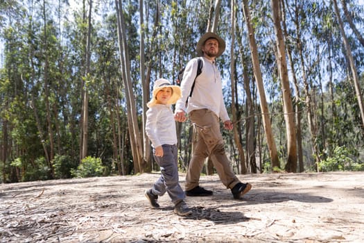 Father and son enjoying a leisurely hike through a sunny forest, holding hands and wearing hats
