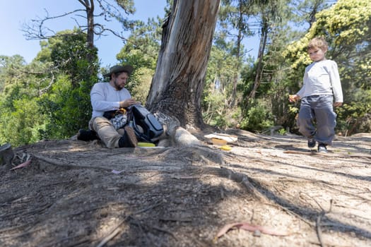 Father and son are having a picnic lunch under a tree in a sunny forest