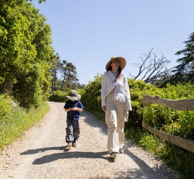 Mother and son wearing hats are walking on a gravel road in nature reserve on a sunny summer day