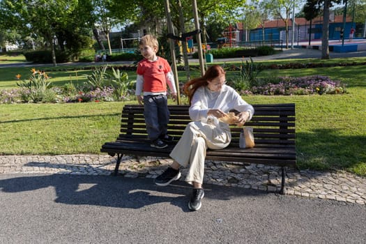 Mother opening a bag of snacks while her son stands on a bench in a park during a sunny spring day
