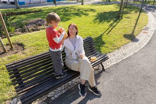 Little boy sharing juice with his grandmother while sitting on a park bench