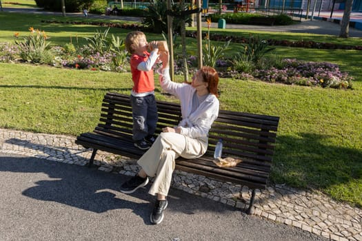 Mother assisting her son drinking juice from a paper carton, while sitting on a bench in a park