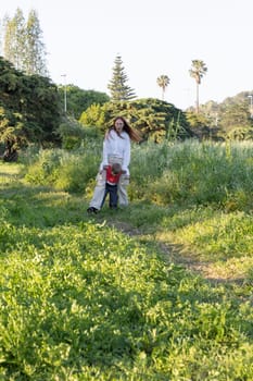 Mother carrying her son while walking across a meadow in a park on a sunny spring day