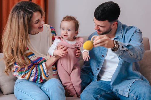 Smiling parents sitting on a sofa while playing with their baby girl using a toy ball. Warm indoor setting ideal for themes like love, family bonding, early development, and parenting.