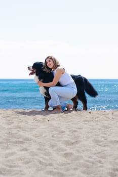 Portrait of a cheerful young woman kneeling beside her dog on a sandy beach. Both look at the camera on a sunny day, enjoying a peaceful moment by the sea with joy, friendship, and nature. Vertical