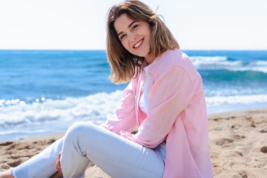 Young caucasian woman on vacation relaxing by the sea, sitting on the beach with a smile. Dressed in casual clothes, enjoying the sunny weather, ocean breeze, and peaceful summer atmosphere.