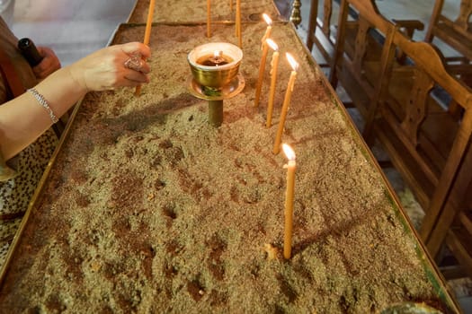 A hand lighting a candle in the Church of Annunciation, a symbolic act of prayer and spiritual devotion, with rows of wooden benches in the background.
