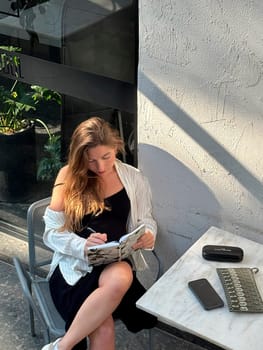A young woman sits at a cafe table, jotting down notes in her journal while enjoying the sunlight in an outdoor space.