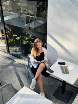 Woman with long hair reads a book at a stylish cafe table surrounded by bright indoor plants in the afternoon light.