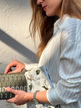 A woman with long hair is holding a patterned pouch and a floral notebook against a light-colored wall, illuminated by soft daylight.