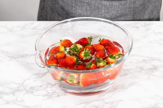 Ripe strawberries are submerged in water within a large glass mixing bowl, a step in washing the fruit to ensure cleanliness and longevity before storage or consumption.