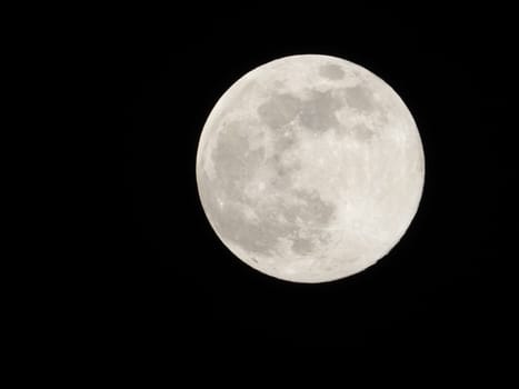 A full moon in the center, in a completely dark sky. Its illuminated surface is highlighted and visible in detail, with several craters and dark spots on a completely black background.