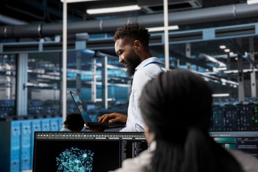 Data center system administrator uses AI visualization tech to oversee automated repetitive tasks. African american man in server farm modifying infrastructure rigs based on data analytics graphs