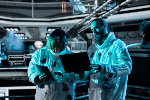 Teamworking astronauts doing brainstorming to confirm suit integrity using laptop. Orbital station colleagues talking, testing gear during routine equipment check using notebook