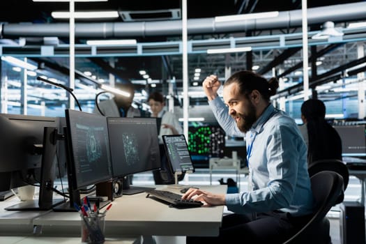 Angry software developer in data center clenching fists while looking at AI visualization interface on monitor. Frustrated technician reacting to artificial intelligence software glitches