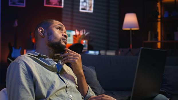 Black teleworker thinking about ways to improve his productivity, looking for new ideas for personal and professional development. Working from home on laptop, networking. Camera A.