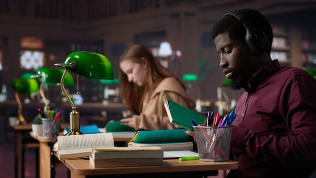 Black guy using study music to help with deep focus for academic success, consulting the campus university database to find reliable sources. Male college student fights for diploma. Camera A.