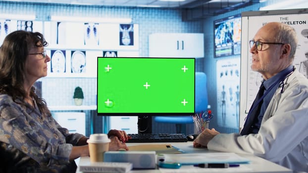 Old general practitioner discusses with female patient next to green screen, offering details from the recovery plan at the medical appointment. Medic consulting woman in cabinet. Camera B.