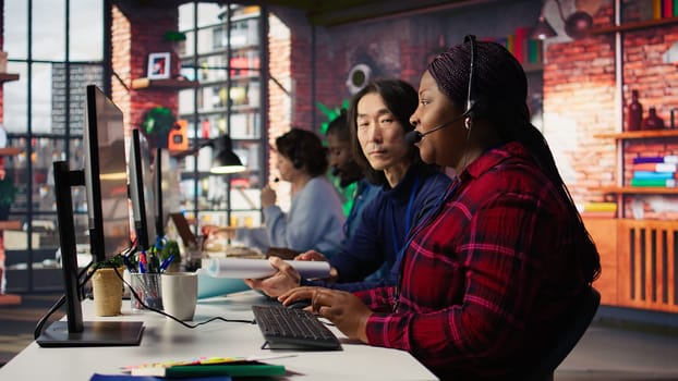 Experienced call center team leader assisting employee and drinking coffee in office. Asian account manager doing teamwork with colleague, enjoying beverage in workplace, camera B