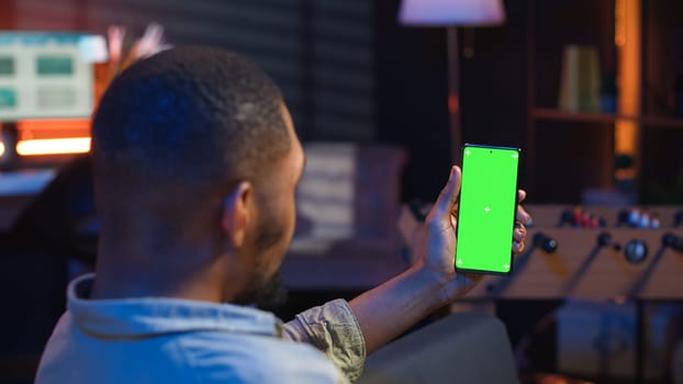 African american young adult laying on the sofa with mockup screen, feeling happy and relaxed with flexible work schedule at home. Freelancer uses phone relaxing in the living room. Camera B.
