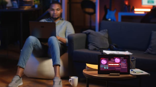 Black IT professional in loft working on cybersecurity with gadgets, tackling hacking attempts, encryption and cyber defense. Screen shows security breach warning, data security risks. Camera B.