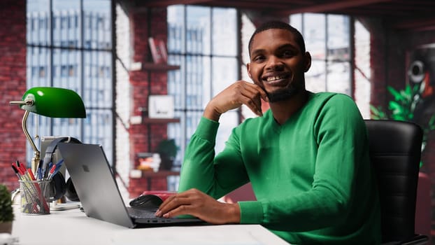 African american remote worker solving his daily freelancing tasks on laptop, working in relaxation from the comfort of his modern loft. Tech savvy black man internet user, multitasking. Camera A.