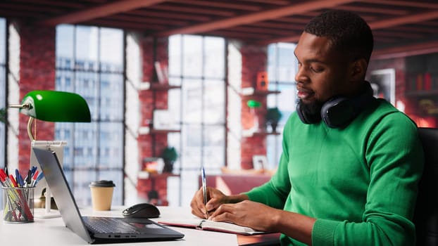Smiling black man taking notes while watching a tutorial presentation, doing online self learning and writing details. Young adult man writes in his textbook notepad. Camera A.